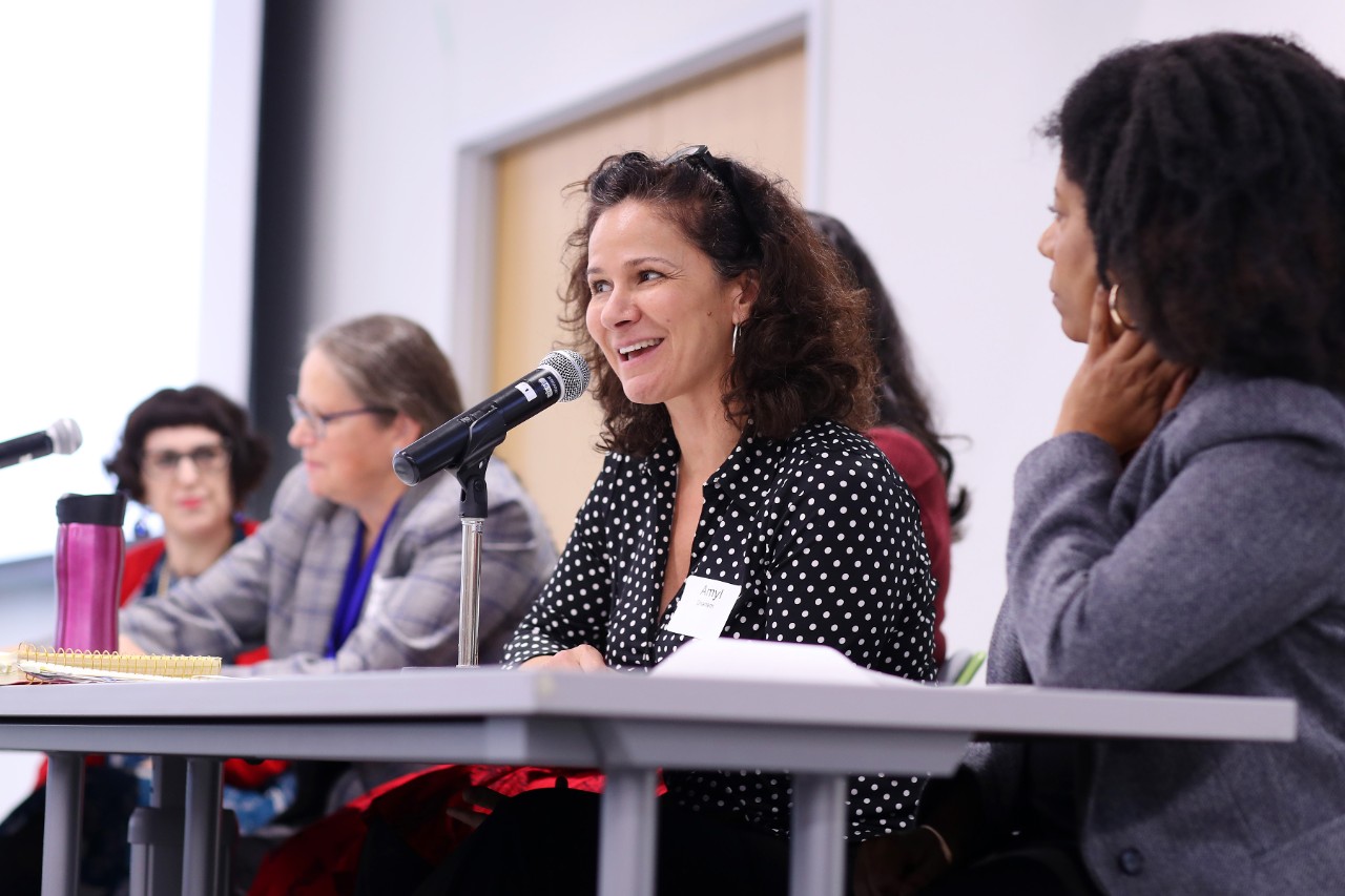 A group of women sit at a long table; one of them is speaking at a microphone. 