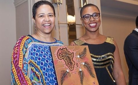 2 Black women stand together smiling. One is holding an artwork featuring an image of Africa surrounded by feathers.