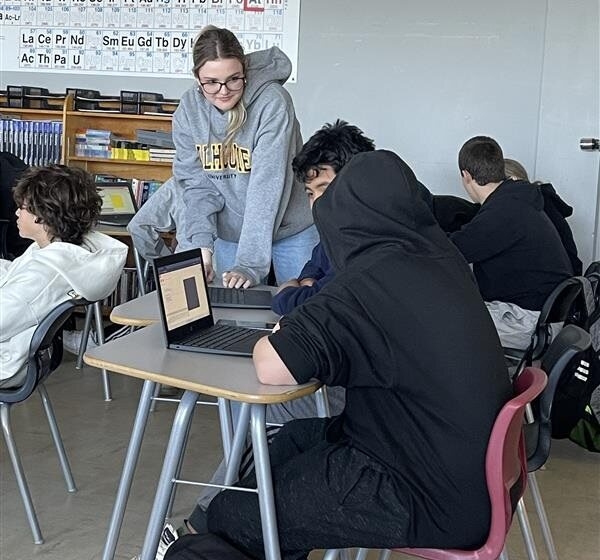 A group of students sit at desks in a classroom with a periodic table on the wall. One student stands and interacts with a classmate working on a laptop.