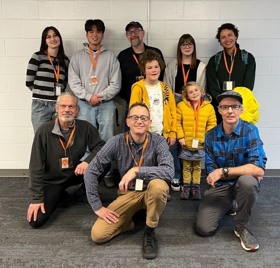 A group of ten people, including adults and children, pose for a photo indoors against a white wall, all wearing orange lanyards.