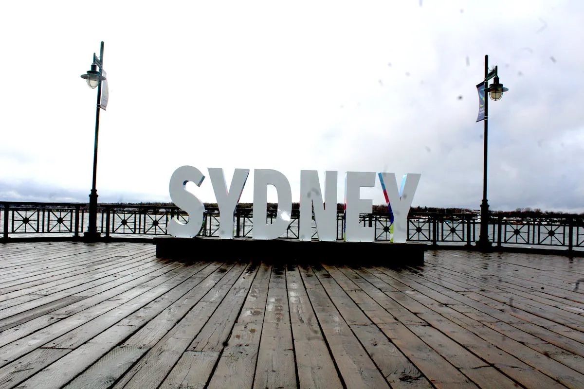 Large 'SYDNEY' sign on a wooden pier with a cloudy sky and two lamp posts in the background