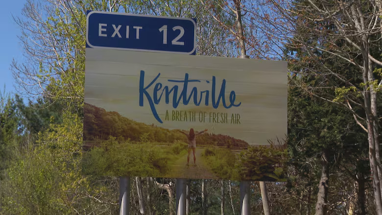 Billboard reading "Kentville, A Breath of Fresh Air" with a person standing on a forested path, arms raised. A highway sign for Exit 12 is above.