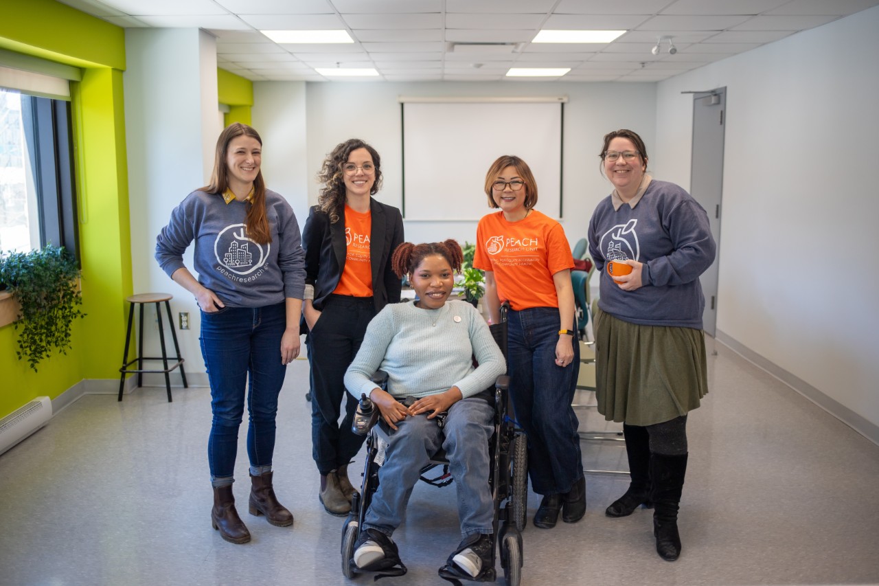 Five individuals wearing casual clothing pose together. The person in the middle sits in a wheelchair.