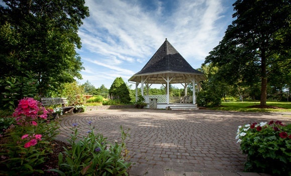 A white gazebo with a shingled roof stands on a circular paved area surrounded by colorful flowers and lush green trees under a blue sky.