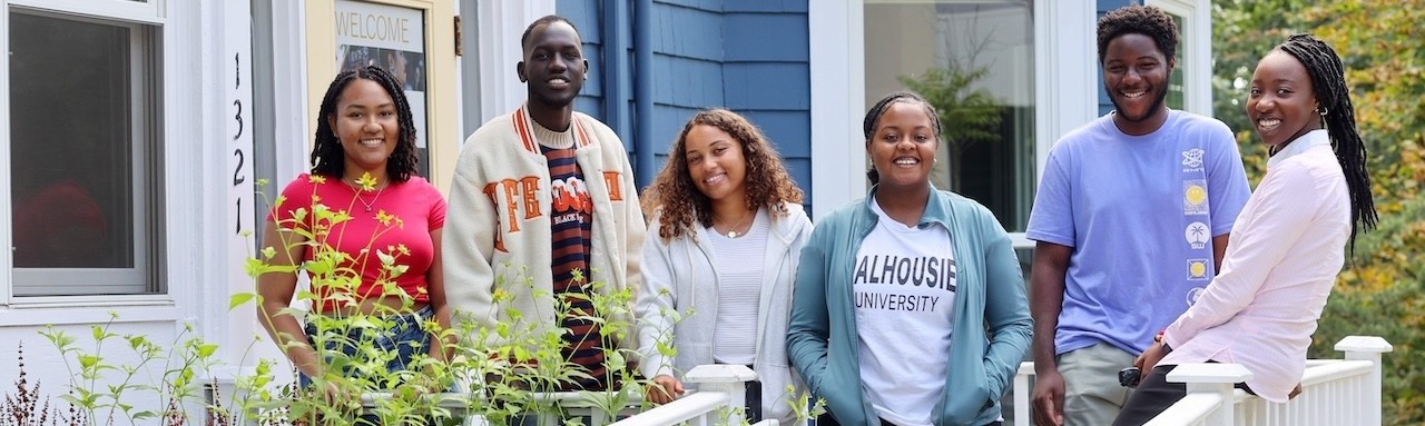 Six people standing and smiling on a porch, with a blue house in the background.