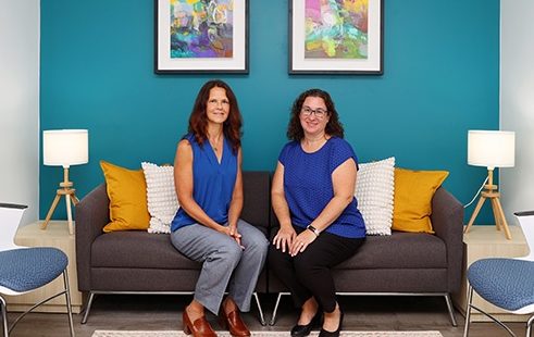 Two women wearing blue shirts sit in front of a bright teal wall.
