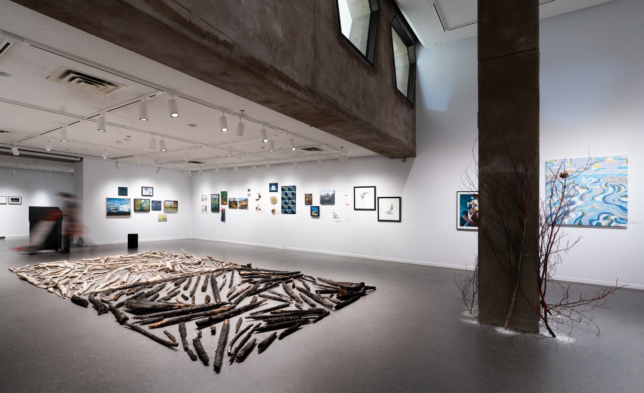  A contemporary art gallery featuring framed artworks on white walls, a floor installation of driftwood and charred branches, and a central concrete column with bare branches. A blurred figure walks past a podium under bright track lighting.