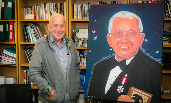 Indigenous elder poses next to a painted portrait of himself.