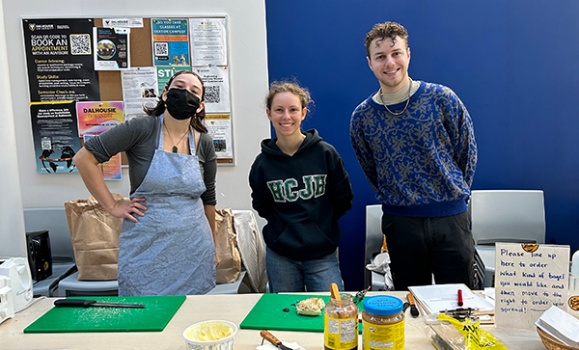 Three students stand at a table with cutting boards, bread, and peanut butter.