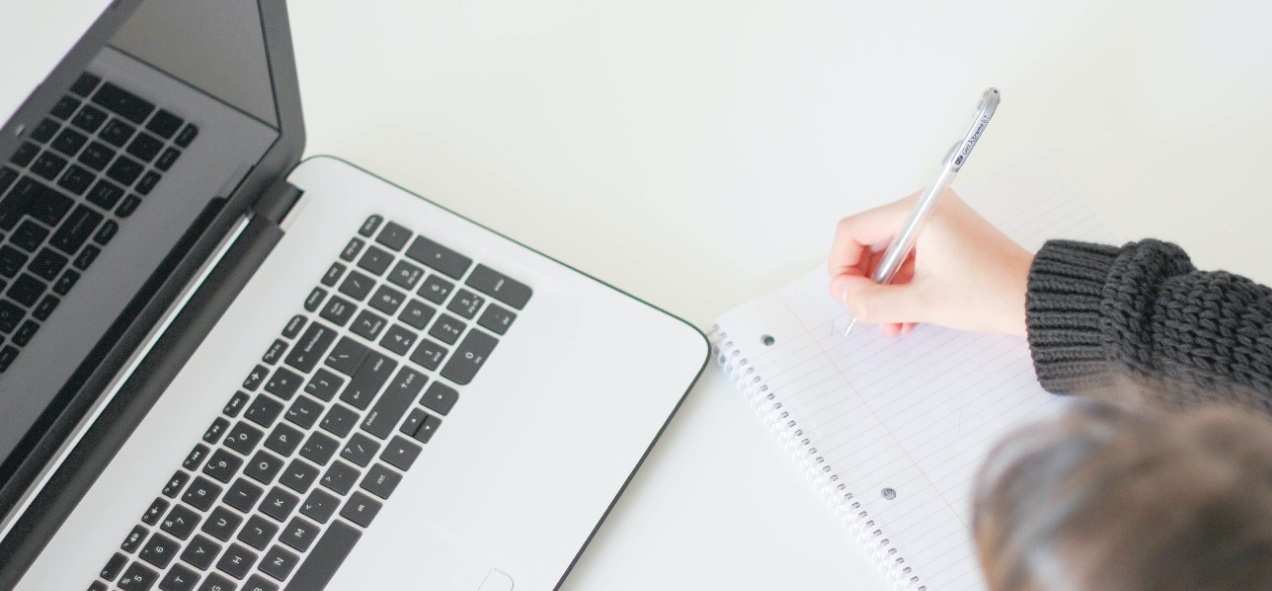 photo of person writing in a notebook with an open laptop next to them