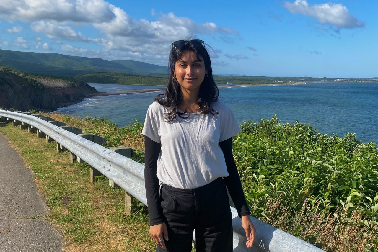 Suha Khan standing outdoors, with the ocean, cliffs, and blue sky in the background.