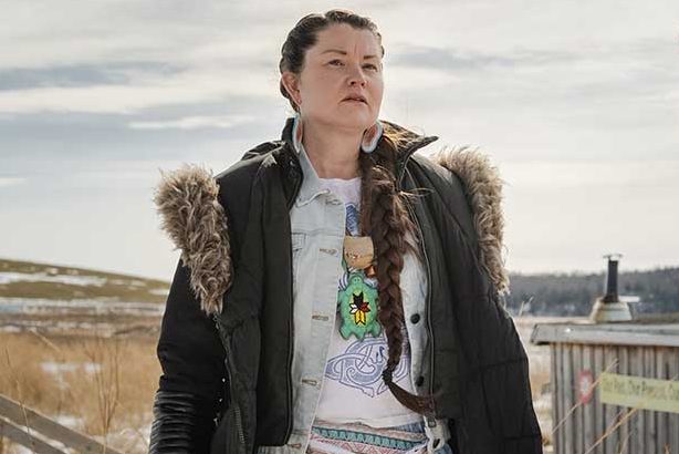 Woman with long dark hair wearing a ribbon skirt and parka standing outside among patches of snow.