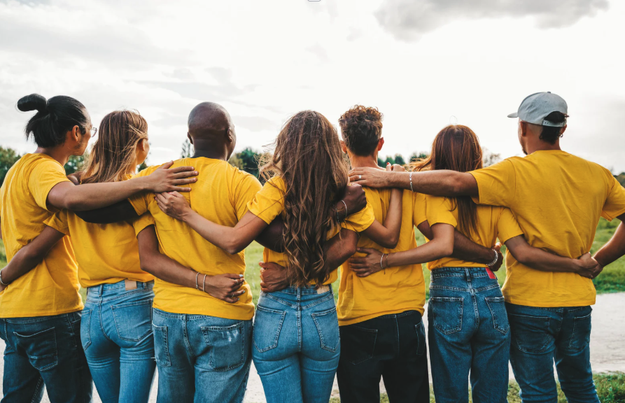 Diverse group of individuals wearing yellow shirts and jeans viewed from behind with their arms around each other.