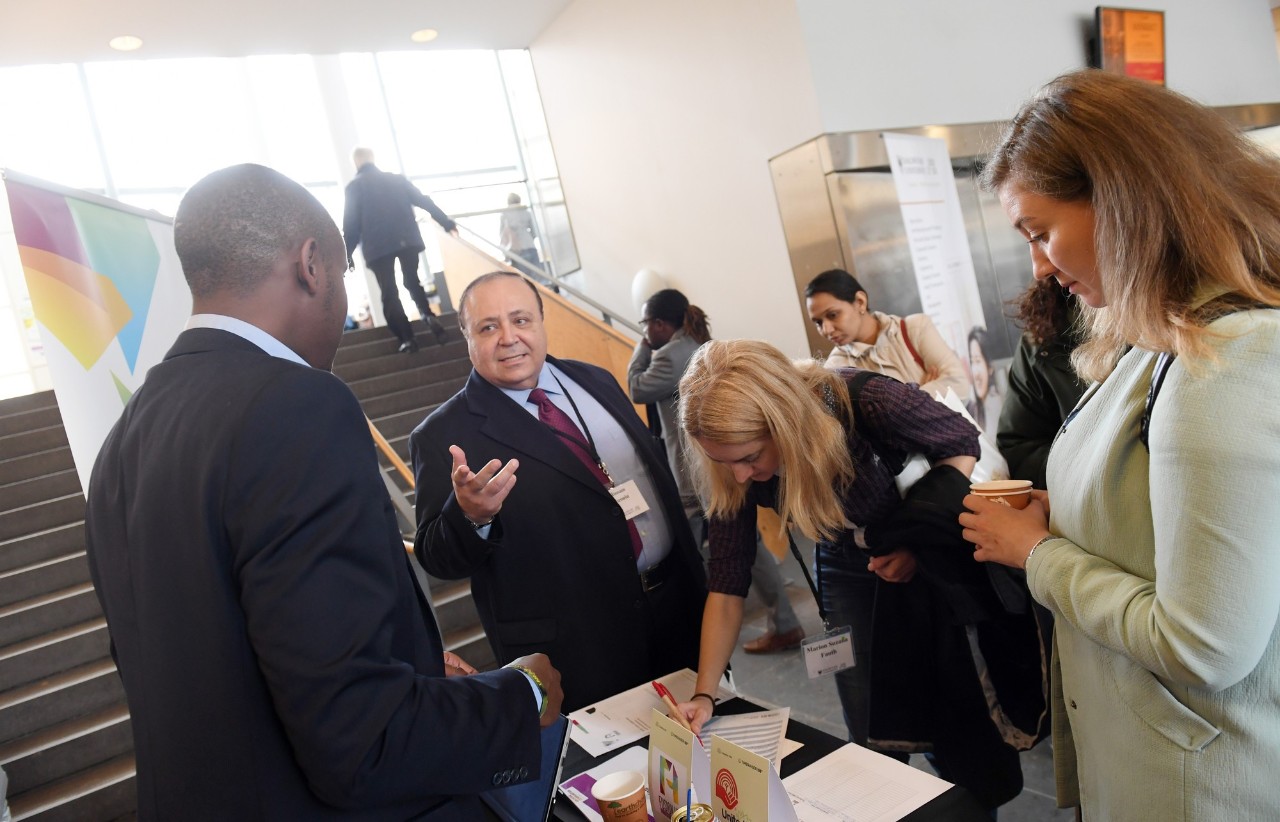 Two men stand talking next to a table, a woman bends over signing a piece of paper and a woman holding a coffee looks on..