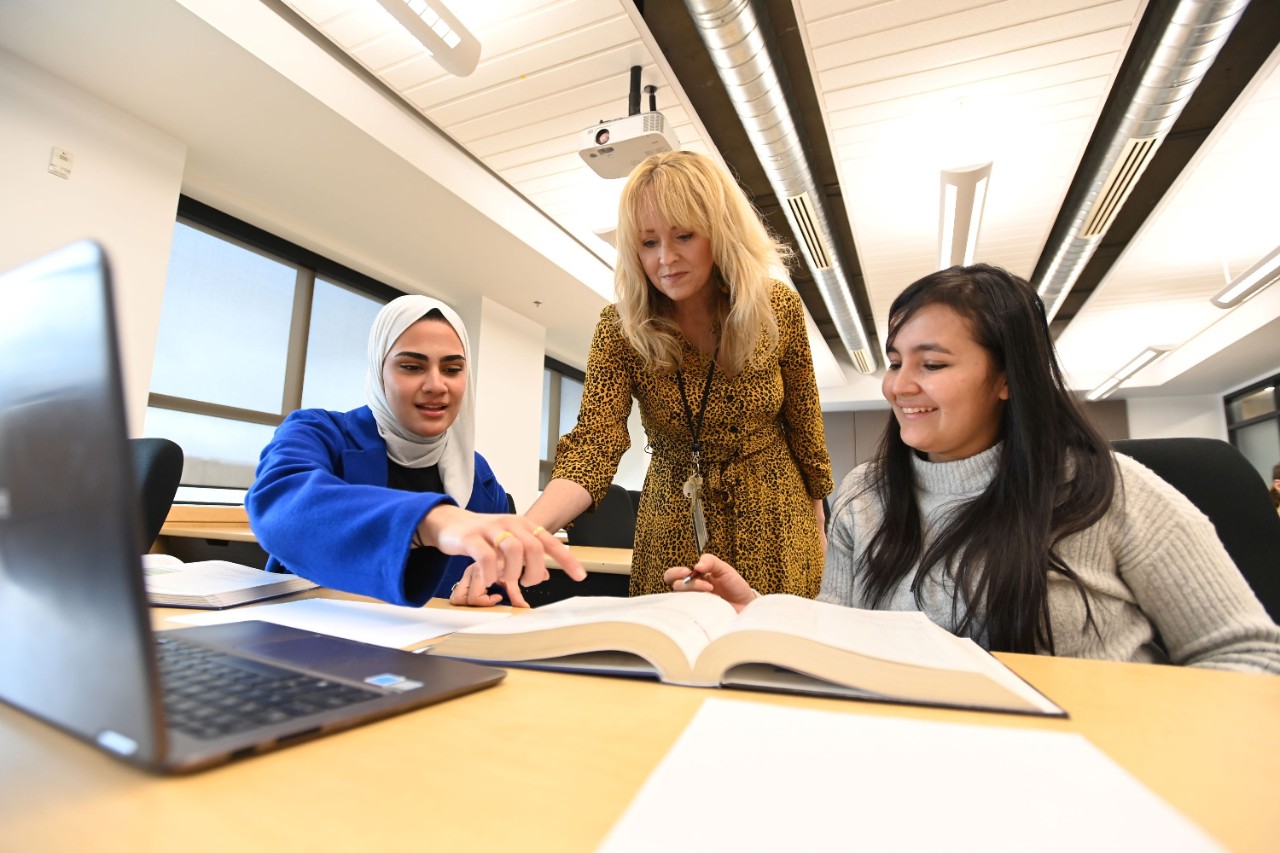 An instructor and two students collaborate over a textbook and laptop at a large table.