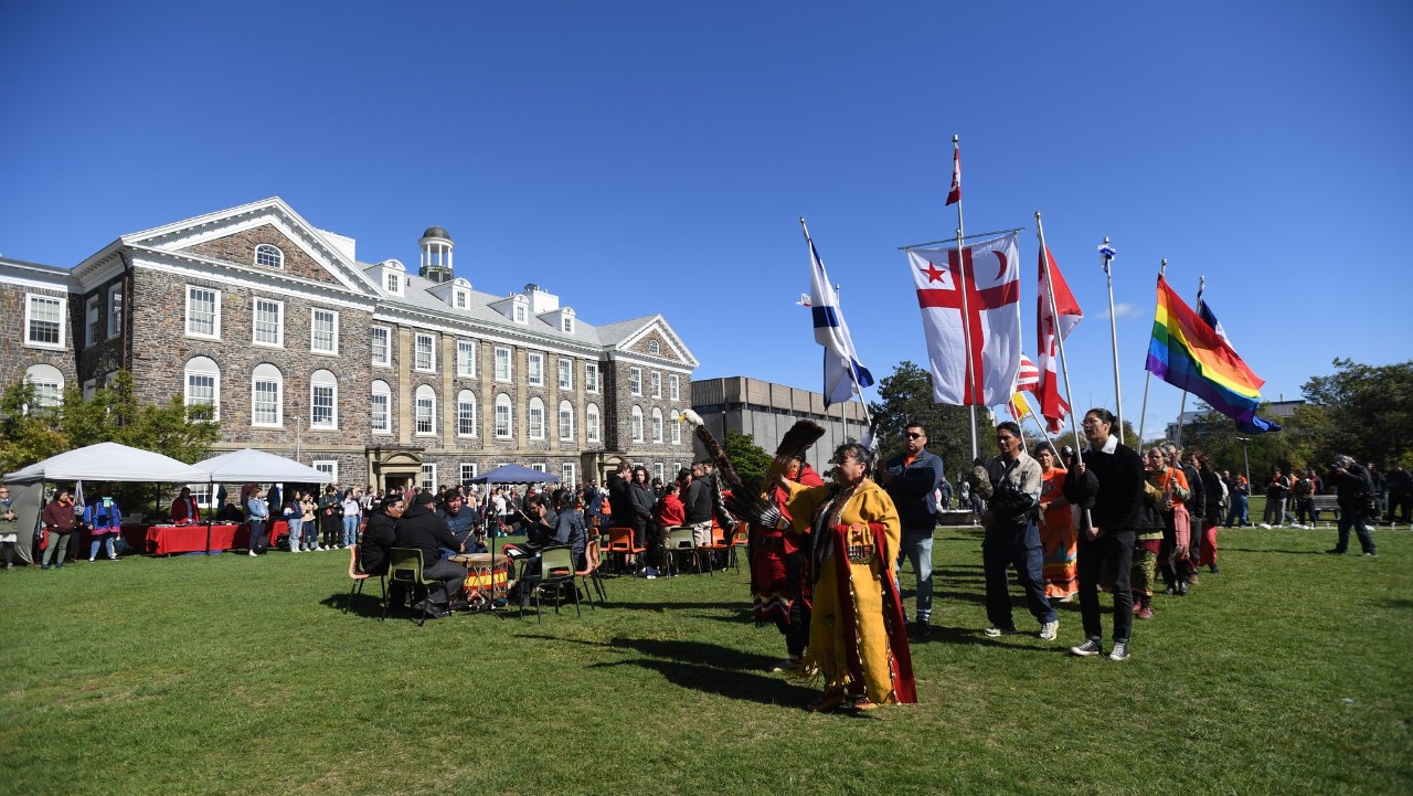 A parade of people in traditional Mi'Kmaq dress and carrying a variety of flags parade through the Studley quad on a sunny day.