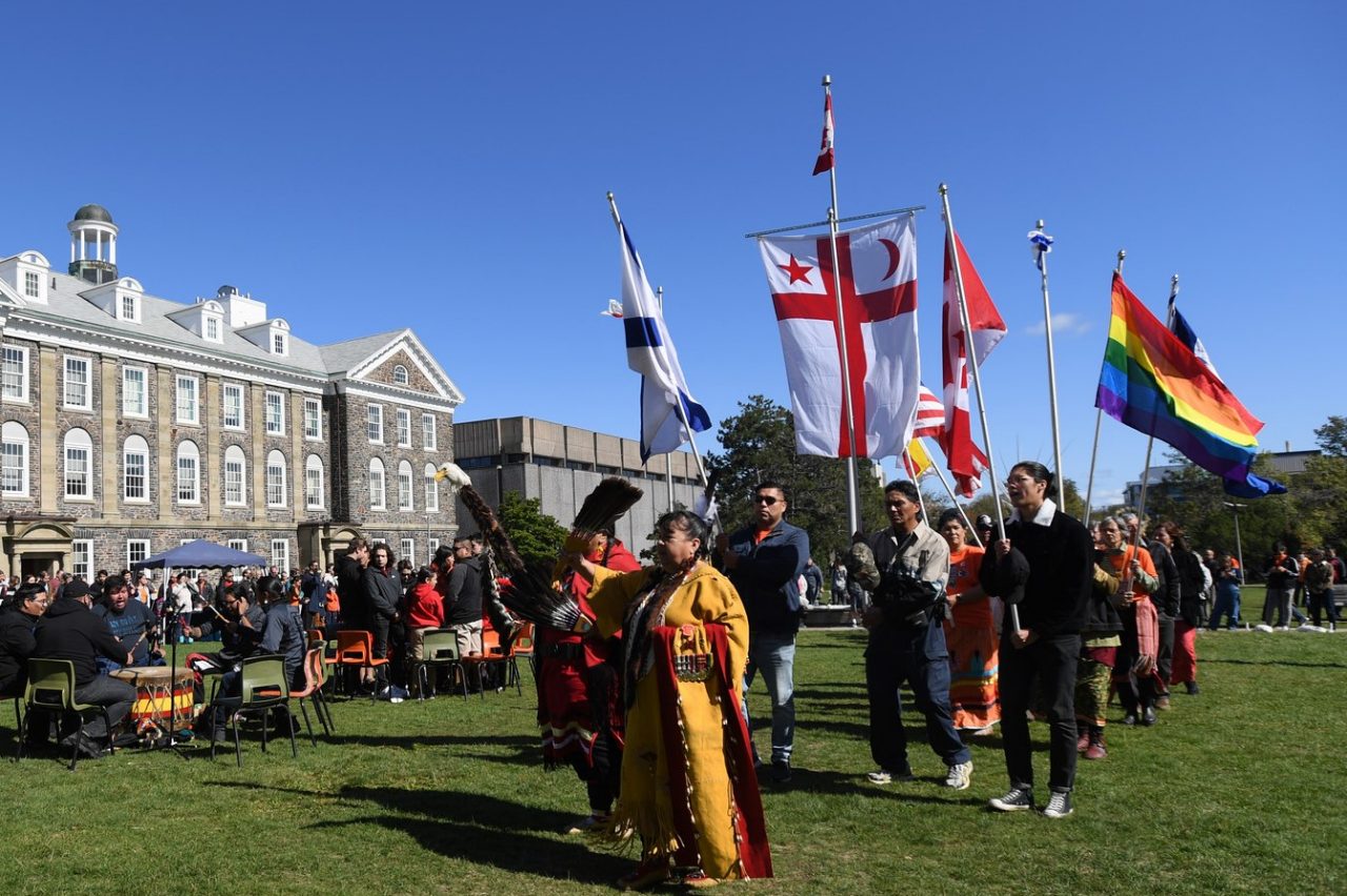 A parade of people in traditional Mi'Kmaq dress and carrying a variety of flags parade through the Studley quad on a sunny day.