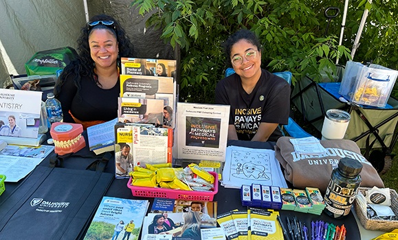 Two Dalhousie students are sitting at a table outside. The table has pamphlets, pens, snacks, and Dalhousie swag.