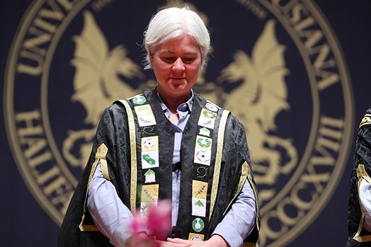 Dalhousie President Kim Brooks standing in front of the convocation banner, wearing a new ceremonial robe with meaningful symbols including fiddleheads, sweetgrass braids, blueberry flowers, the African Nova Scotian Affairs Mayflower logo, and lavender.
