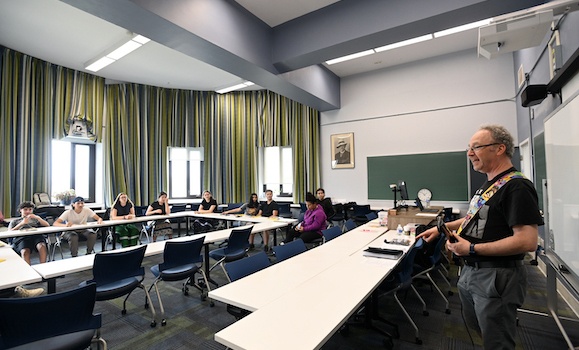 Dr. Jason Brown stands at the front of a classroom, playing a guitar. 