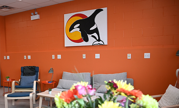 A room with orange walls, a portrait of a killer whale and sun, four chairs, and a bouquet of flowers.
