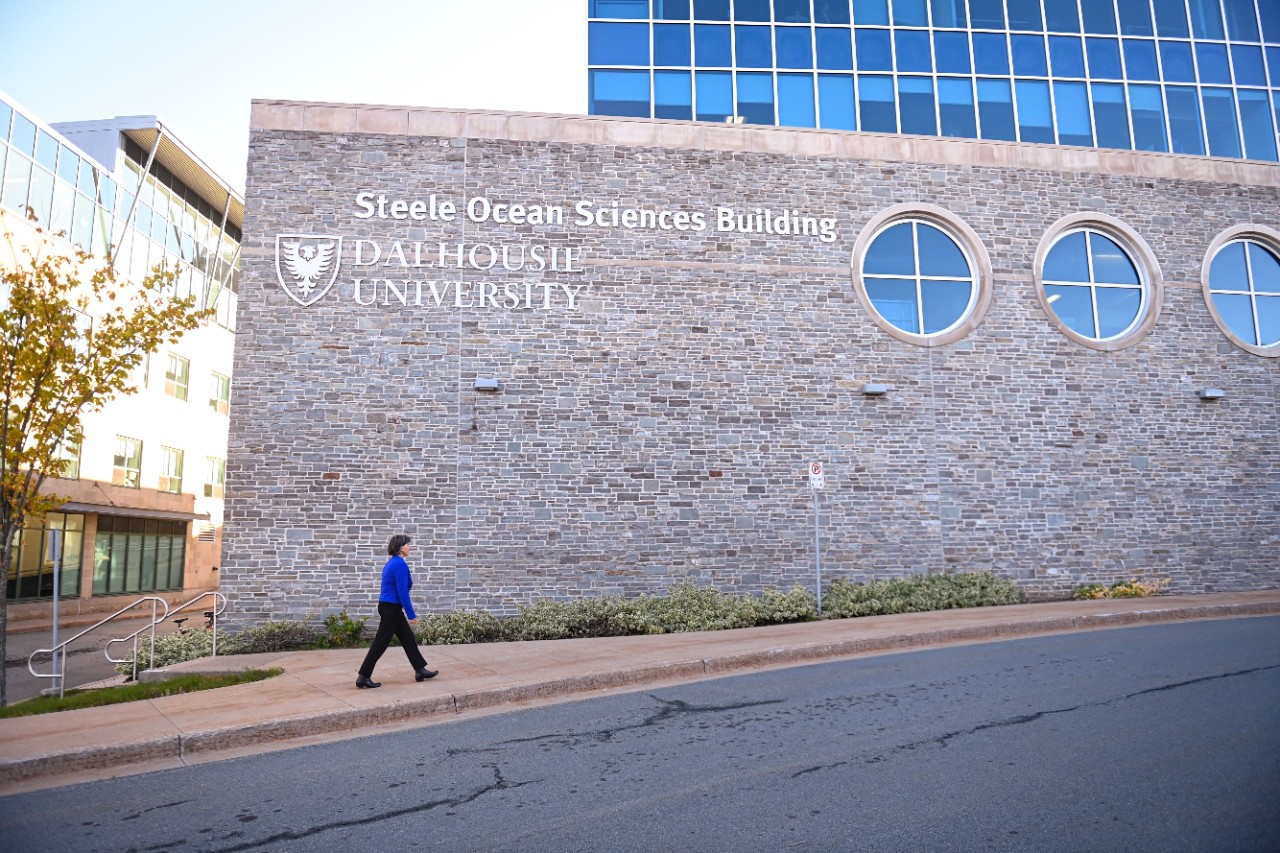 A woman wearing a blue jacket walks in front of the stone Steele Ocean Sciences Building at Dalhousie University. 
