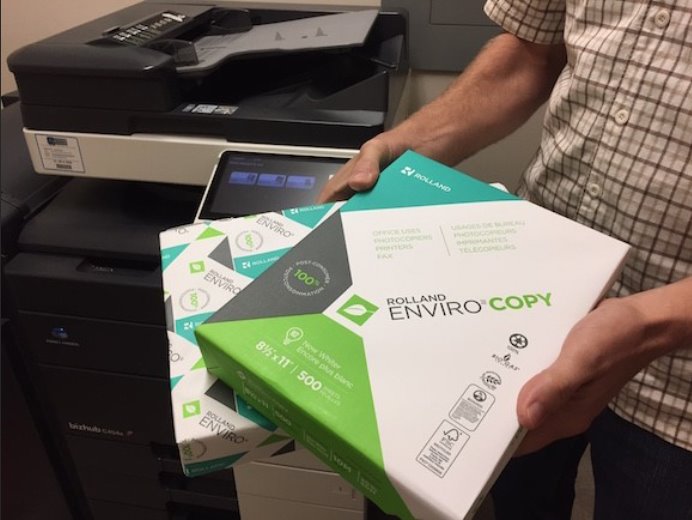 A person holding packages of environmentally friendly printing paper while standing beside a printer.