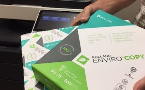 A person holding packages of environmentally friendly printing paper while standing beside a printer.