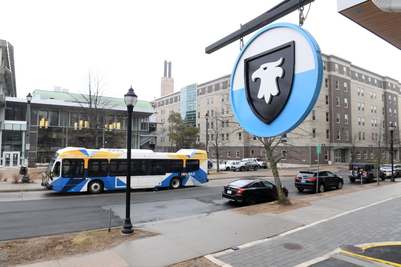 A blue, black, and white Dal logo sign hangs outside a building, with a Halifax Transit bus in the background. 