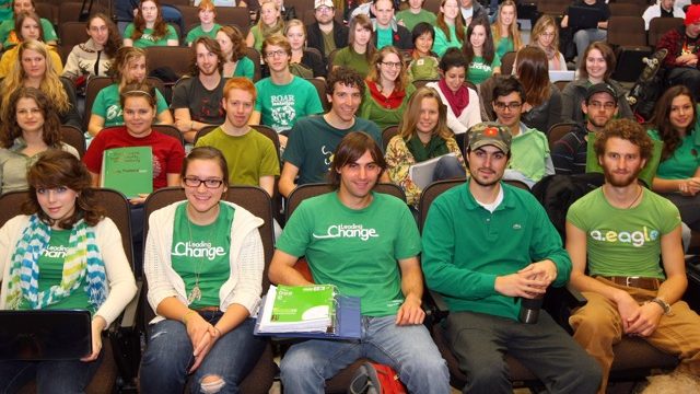 A group of students all wearing green shirts sit in a lecture hall.