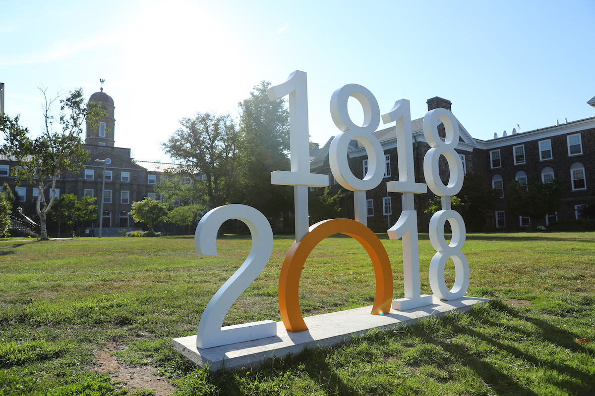 A large sign reading 1818-2018 stands on a green lawn in front of Dalhousie University's Henry Hicks building.