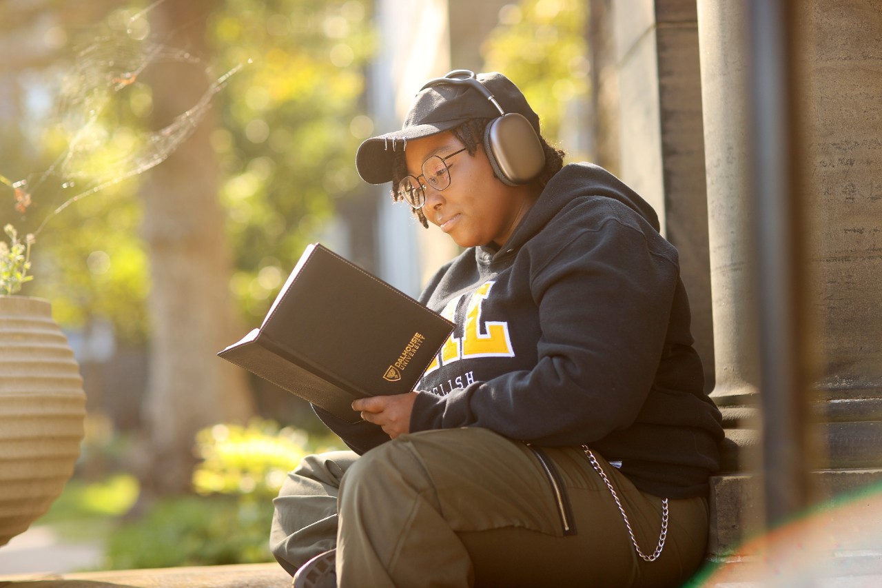 A student wearing headphones and a Dal sweatshirt sits outside reading a book in the sun. 