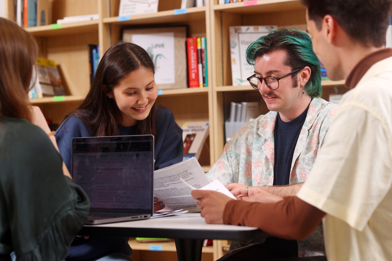 A student with pink dyed hair, wearing a tie-dyed shirt and cardigan, sits in a chair with her legs over the armrest while writing in a notebook.