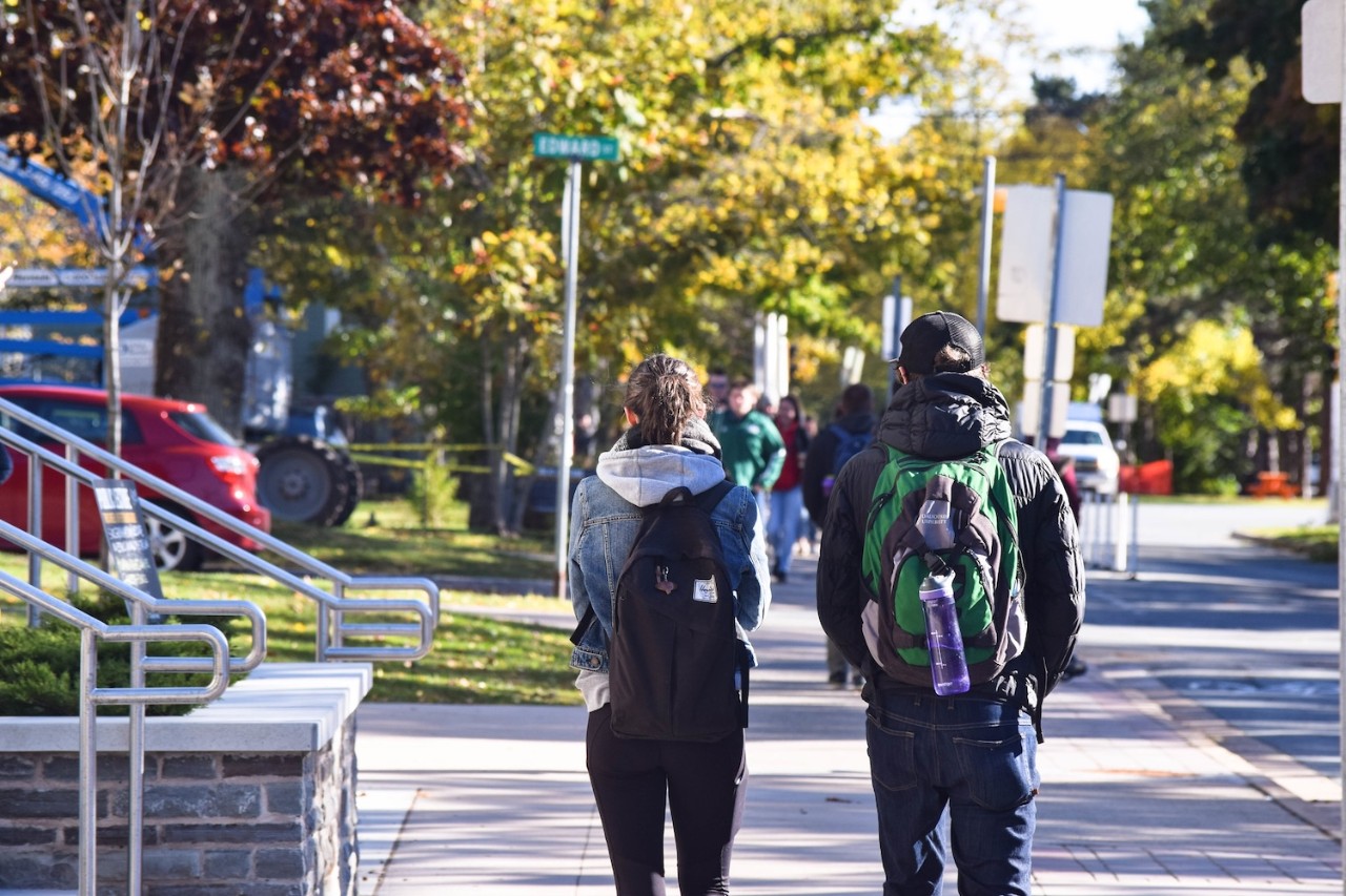 Two students wearing backpacks walk along the sidewalk with trees in the background.