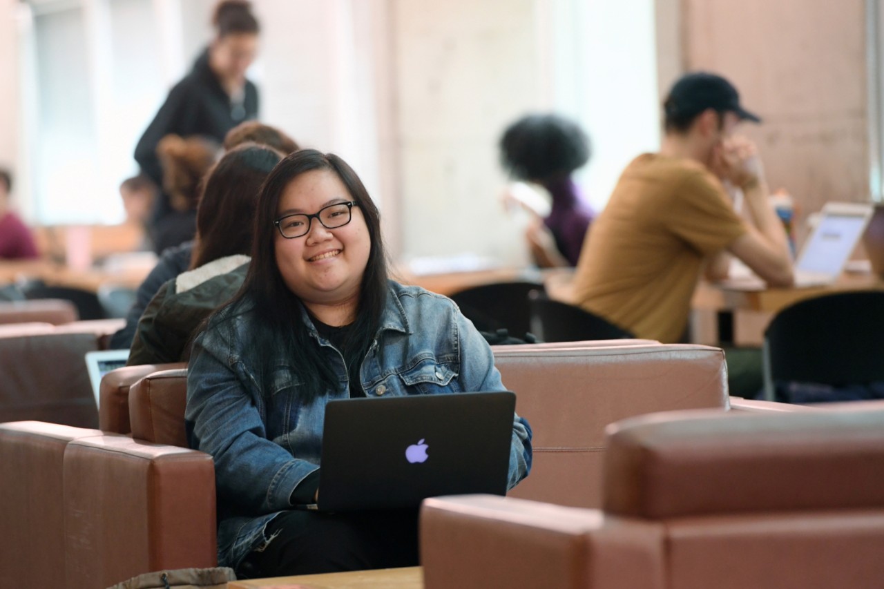 Alicia Wong is seated with her open laptop in a learning commons setting with others on laptops in the background.