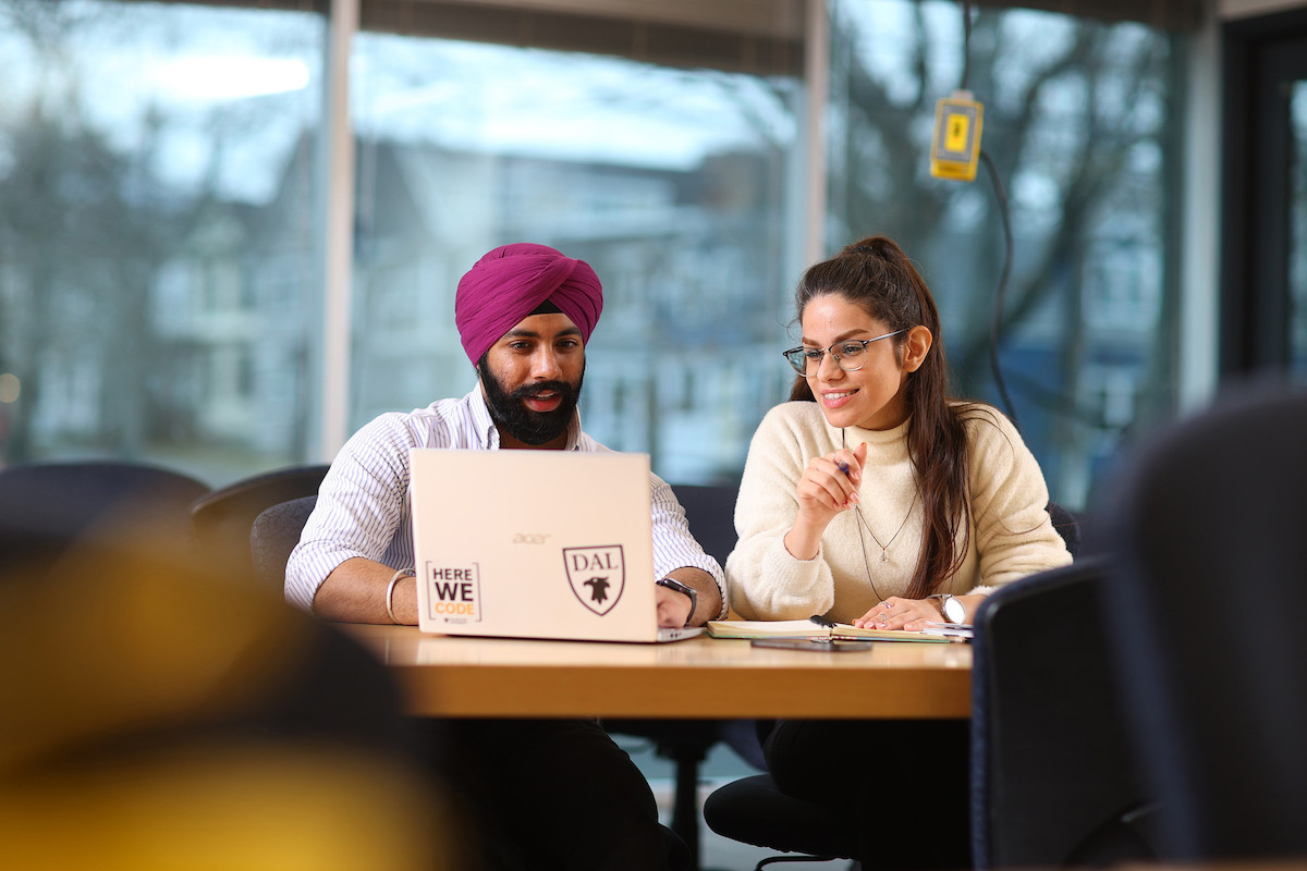 Two students sit together at a table. One is using a laptop.