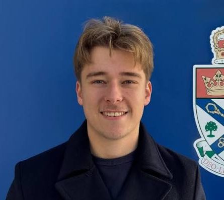 Student with short blonde hair with a flag and crest in the background.
