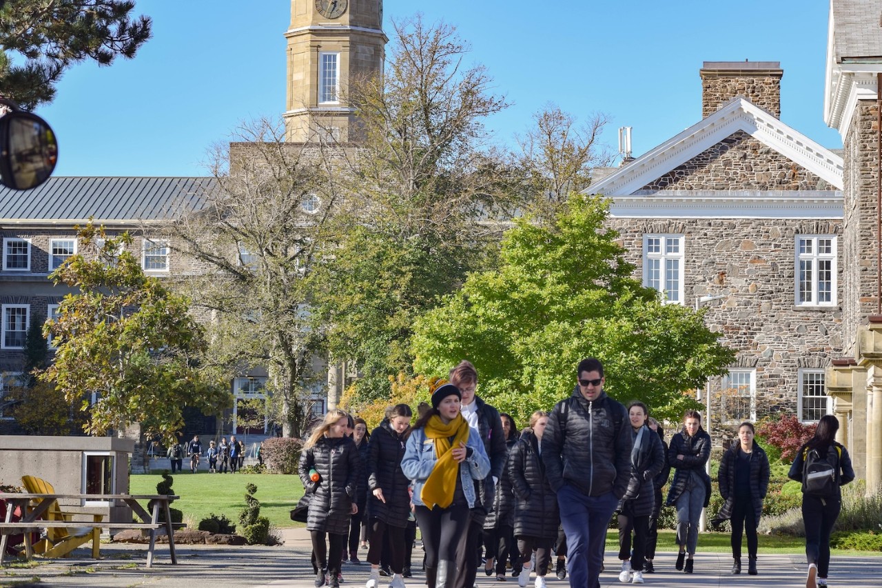 A large group of students walk through the Studley Quad with the Hicks Building behind them.