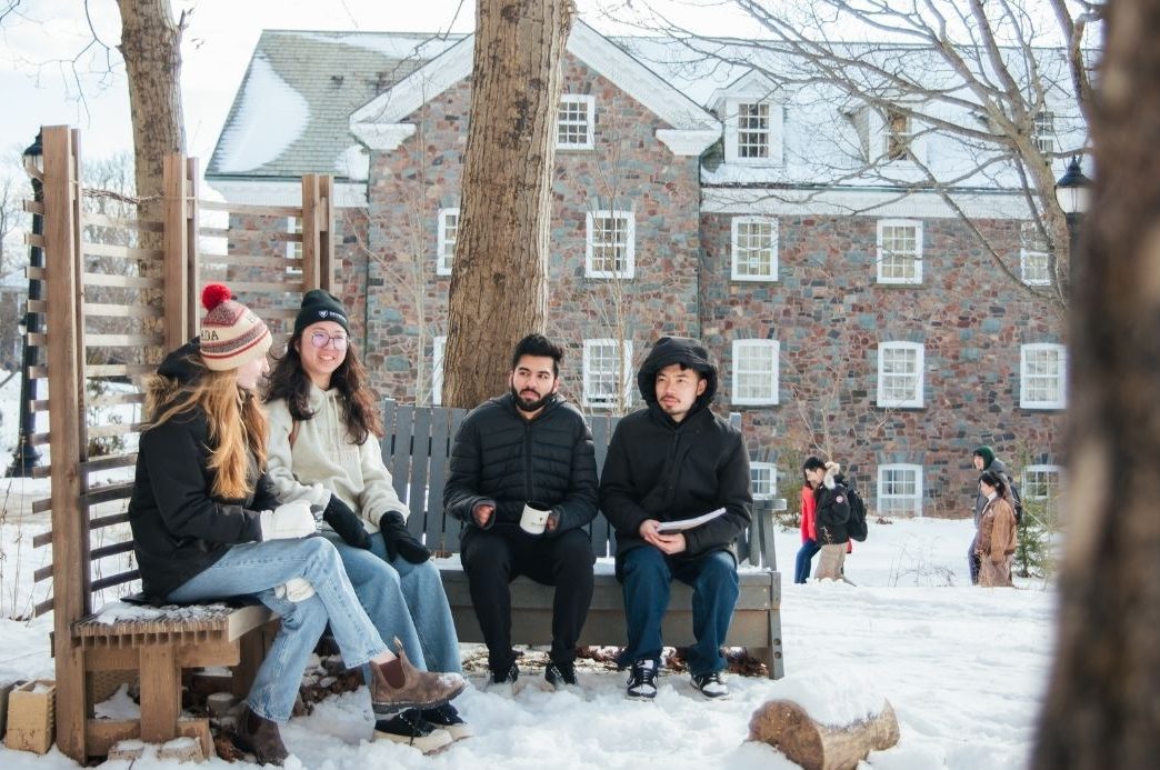 Four students sitting on benches outdoors on campus in winter with snow on the ground.