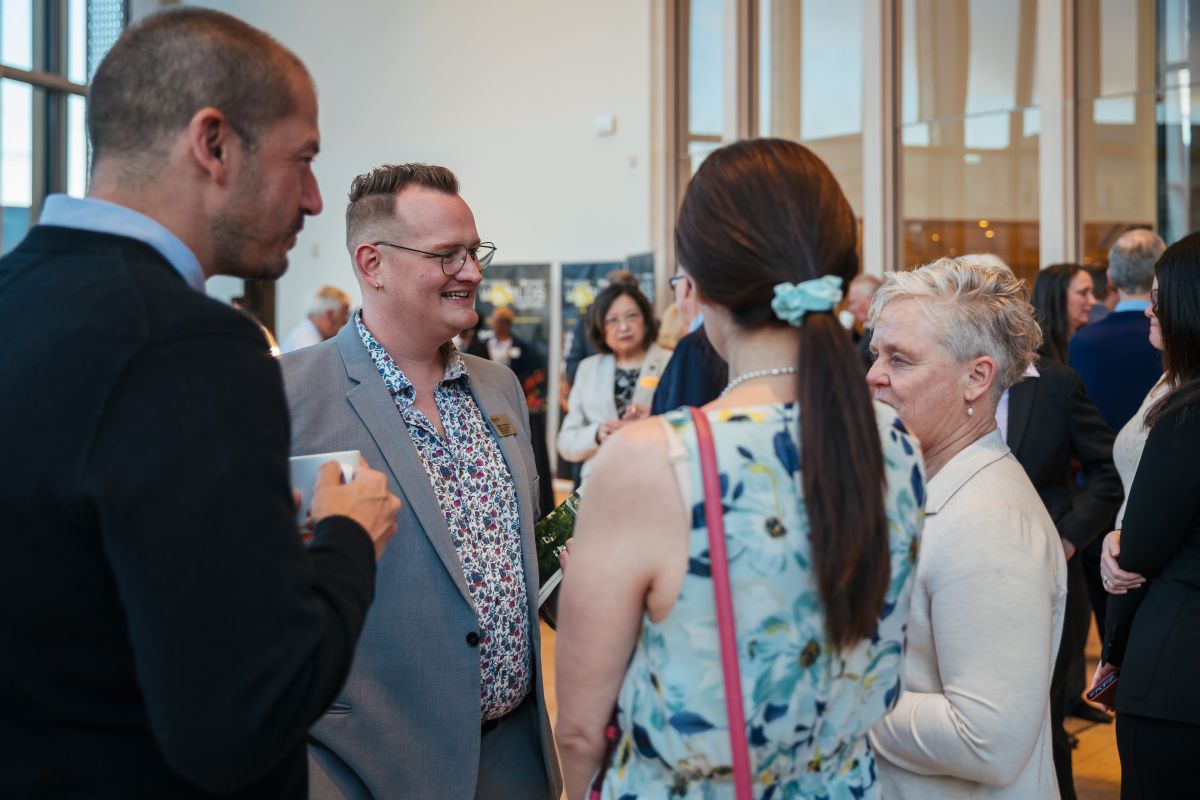 Two men and two women stand in a circle mingling at an event with more attendees in the background.