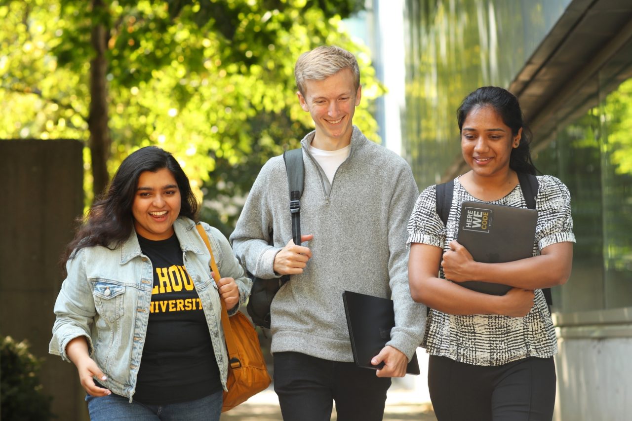 A group of three students walking down a sidewalk with backpacks and laptops.