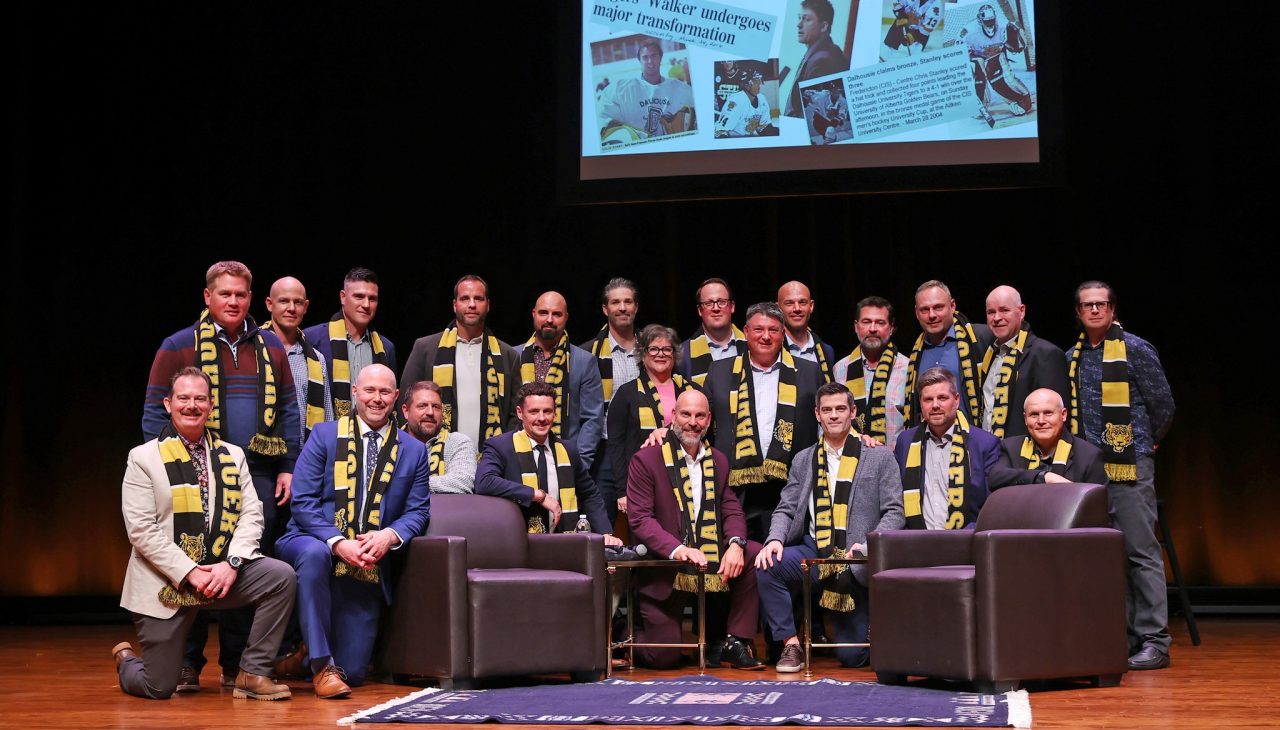 A group of former student-athletes wearing black and gold scarves pose on stage at an event.
