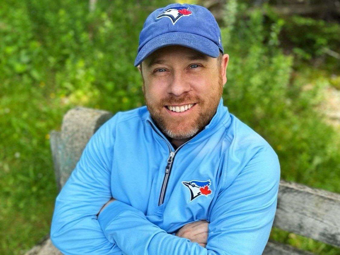  Josh Goreham in Blue Jays cap and pullover is sitting on a bench outside smiling with arms crossed.