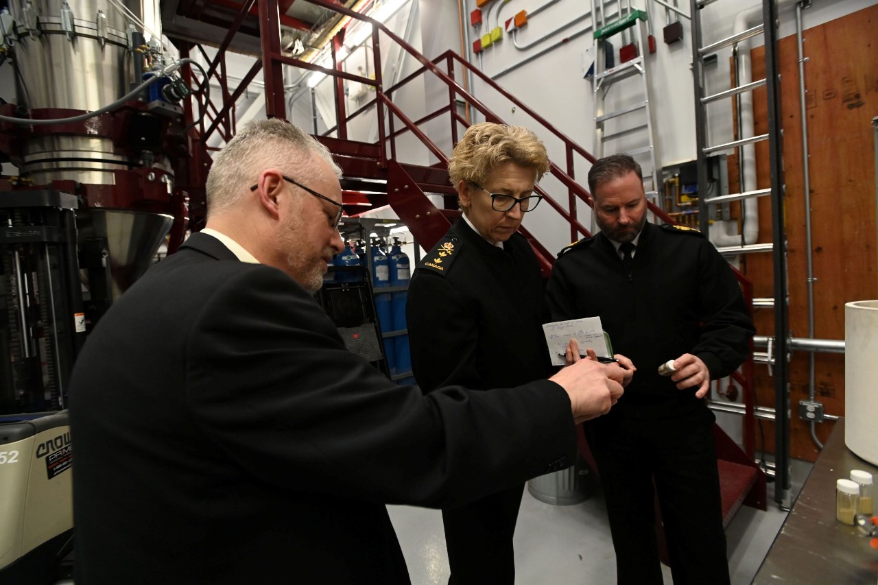 Three people in formal uniforms stand together inside an industrial laboratory, examining a small object and a notepad. Behind them are large metal machines, pipes, and a red metal staircase.