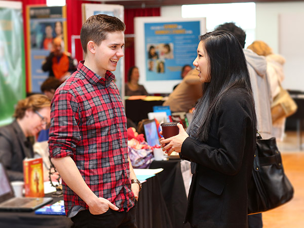 Two people are in conversation at a career fair with tables set up in the background and banners.  