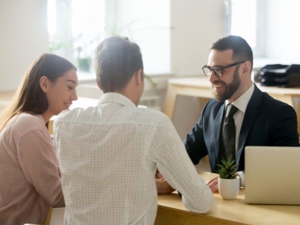 Two people sit across a desk from a male-presenting person who is dressed in a suit and tie in an office setting.