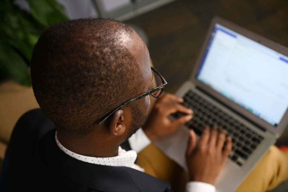 A man sits and types on a laptop. The perspective is from behind him with the screen visible. He is dressed in business casual attire. 