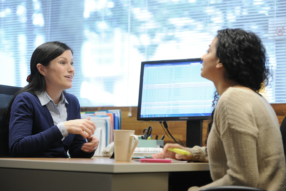 Two women conversing over a desk with a computer screen in the background.