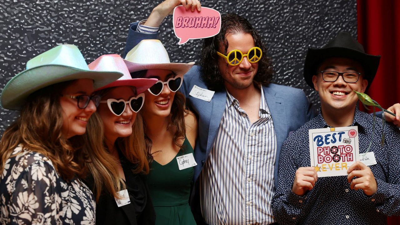 Event attendees pose wearing heart-shaped glasses, cowboy hats, other props and nametags.
