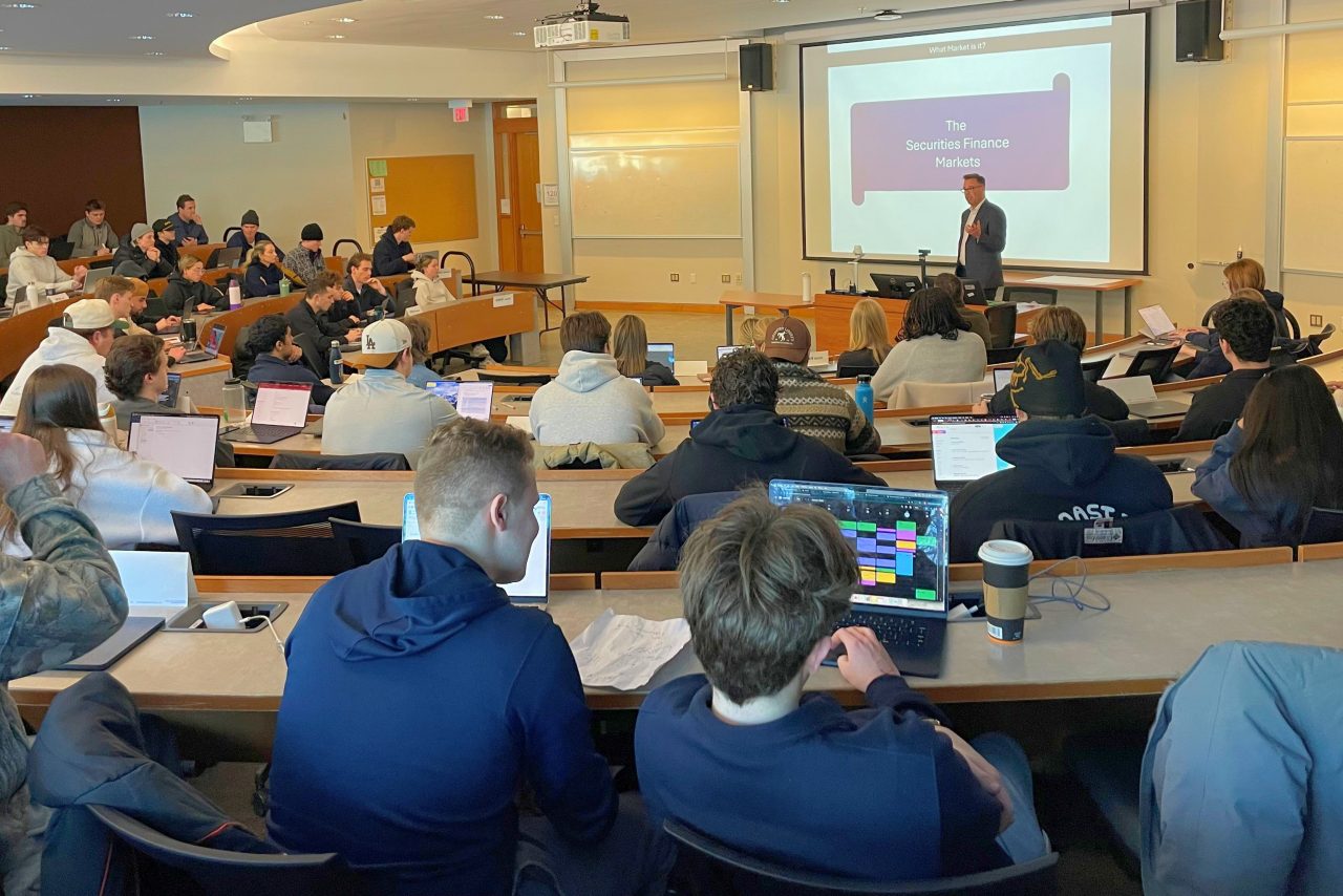 Students sitting in a lecture theatre working on laptops and focused on a person presenting and a screen at the front of the classroom.